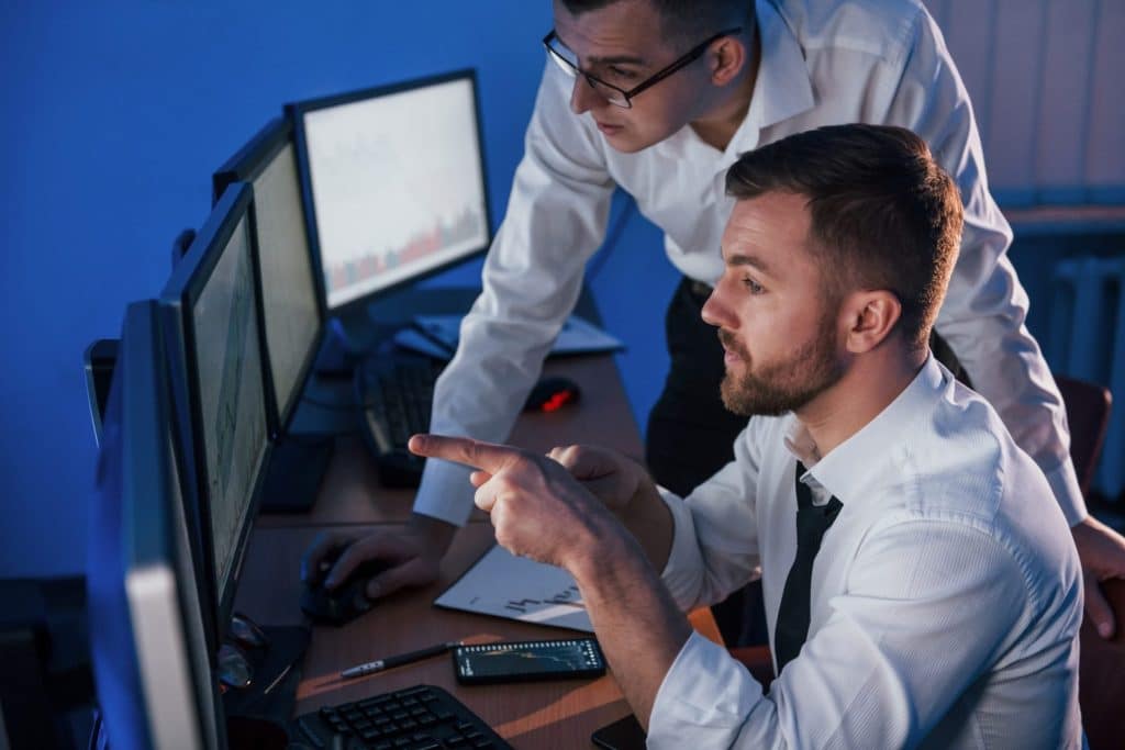 Photo of two men looking at computer screens illustrates blog "CMMC Continuous Monitoring: What Is It and Why Is It Important?"