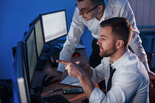 Photo of two men looking at computer screens illustrates blog "CMMC Continuous Monitoring: What Is It and Why Is It Important?"