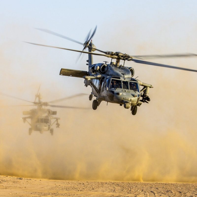 Two military attack helicopters descending on a dirt runway.