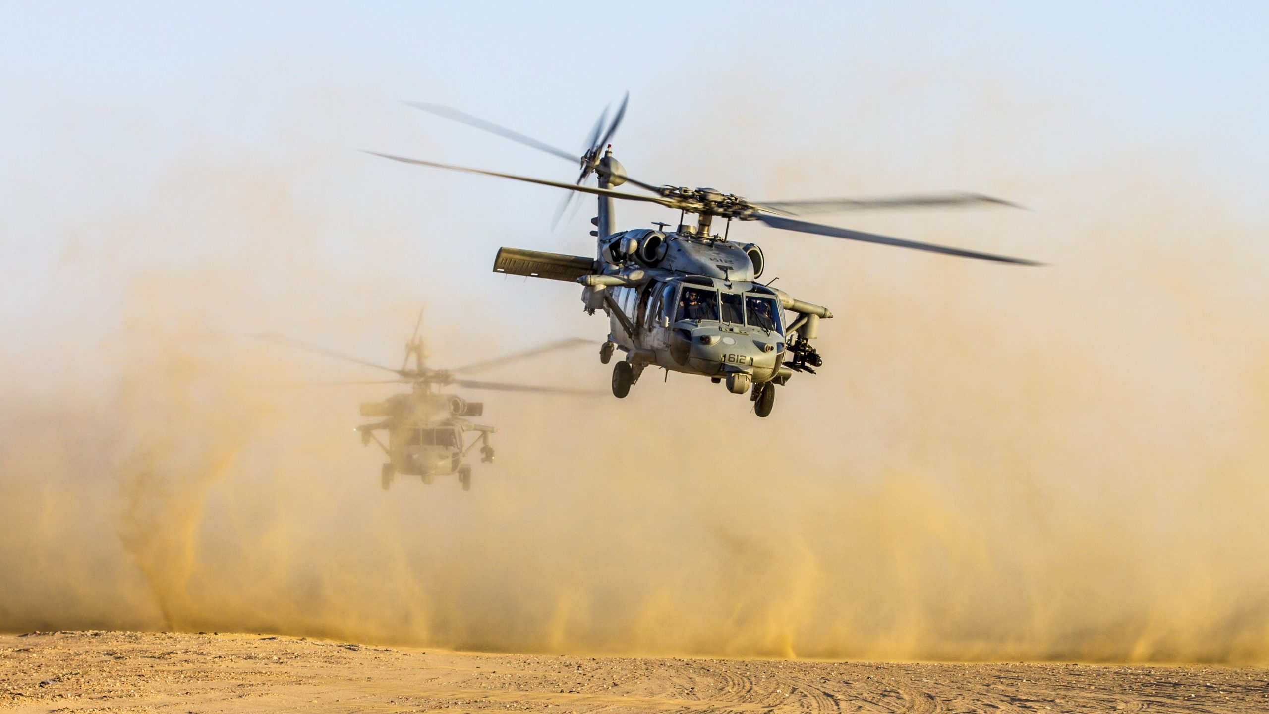 Two military attack helicopters descending on a dirt runway.